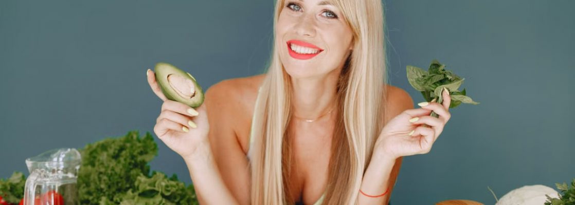 Blonde woman promotes healthy eating with fresh vegetables and fruits on display.